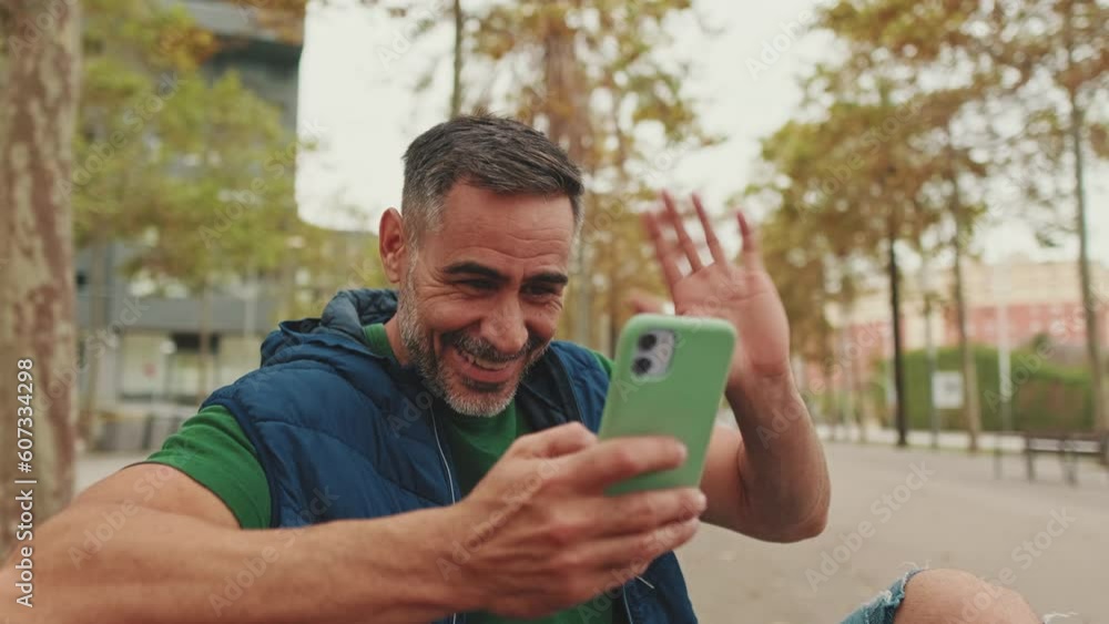 Middle-aged man sitting on bench talking on video call from mobile phone Middle-aged man sitting on bench talking on video call from mobile phone
