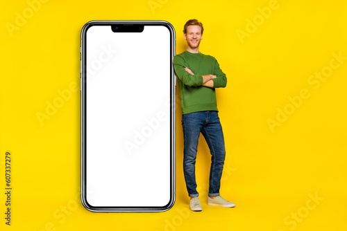 Full length photo of charming handsome guy dressed sweater arms folded modern device empty space isolated yellow color background