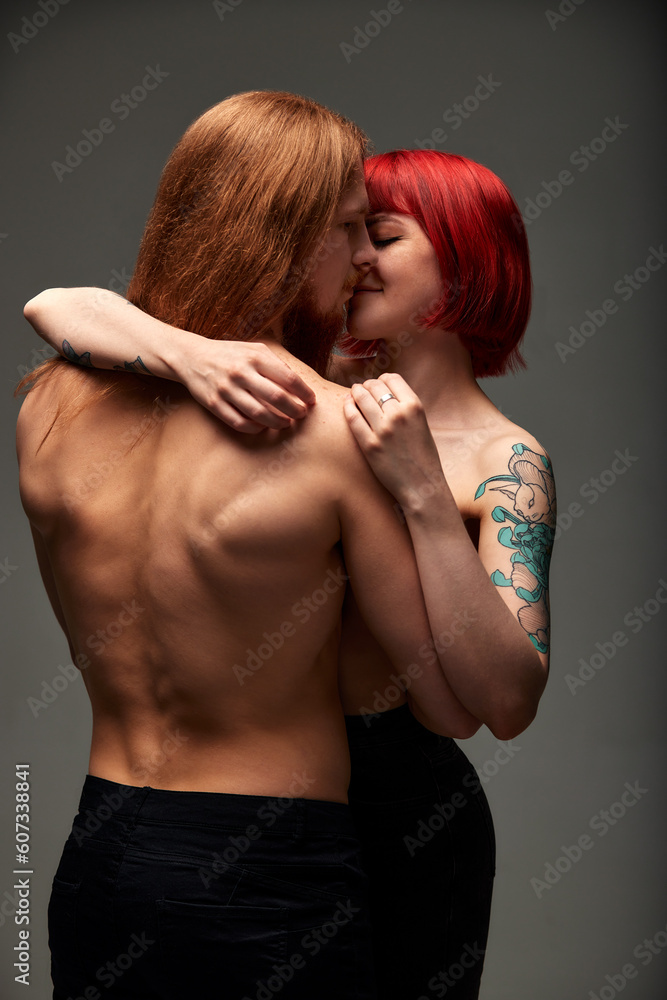 A young attractive woman with bright ruby hair and a tattoo hugs and kisses a man with long hair. Half-naked lovers in a passionate kiss. Half-length studio shot, dark background. A young attractive woman with bright ruby hair and a tattoo hugs and kisses a man with long hair. Half-naked lovers in a passionate kiss. Half-length studio shot, dark background.