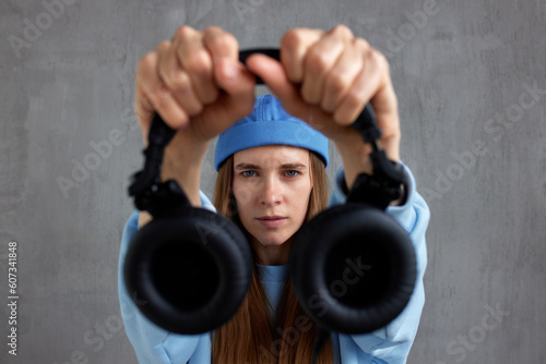 A young pretty long-haired DJ girl in a blue sweater and a funny blue hat holds black headphones in outstretched hands. Studio shot, gray background. photo