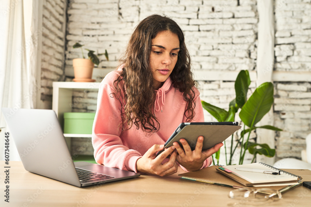 A young woman, focused and productive, working at her home desk with a laptop and a tablet, utilizing technology efficiently. A young woman, focused and productive, working at her home desk with a laptop and a tablet, utilizing technology efficiently.