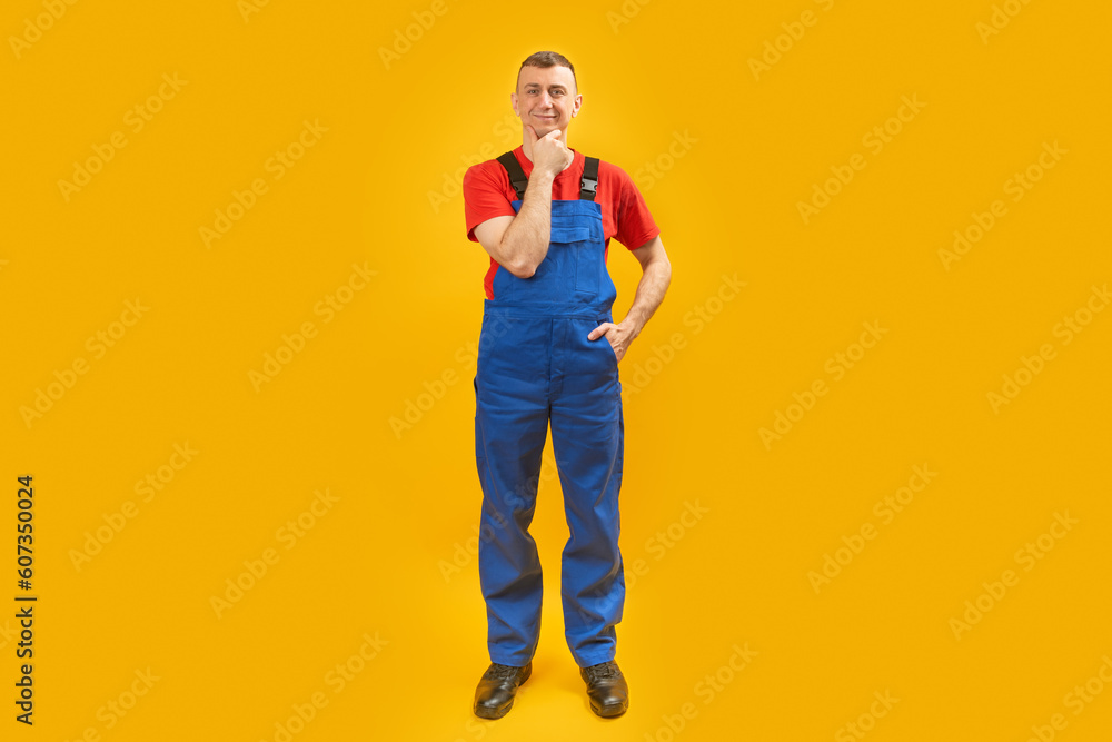 Full-length portrait of builder or worker man in blue jumpsuit put his hand on his chin and thinks. Isolated on yellow background. Copy space. Full-length portrait of builder or worker man in blue jumpsuit put his hand on his chin and thinks. Isolated on yellow background. Copy space.