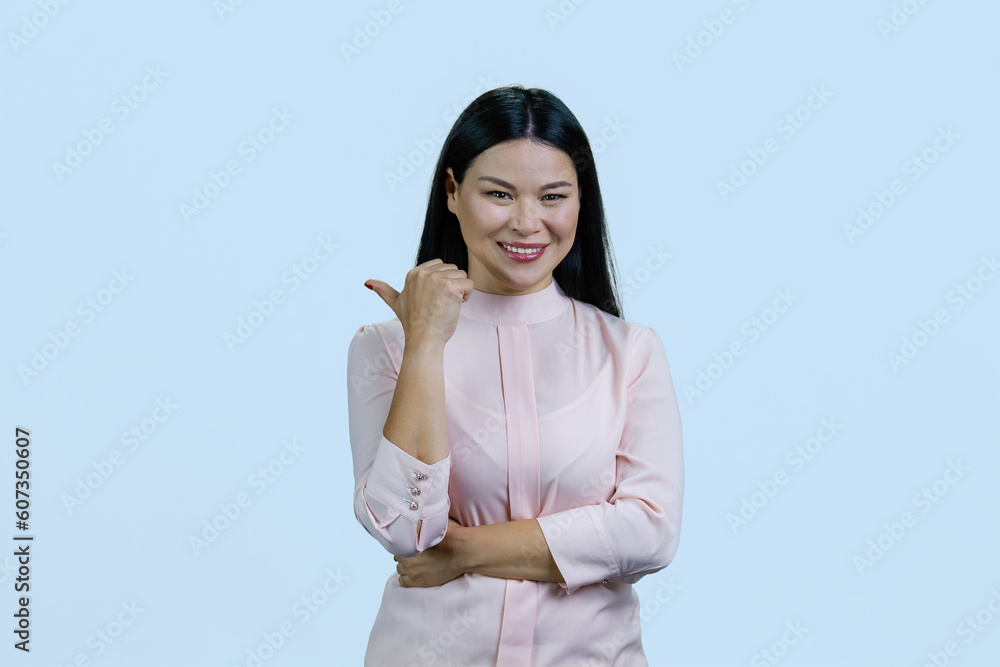 Portrait of happy cheerful asian woman is pointing back with her thumb. Isolated on pastel blue background. Portrait of happy cheerful asian woman is pointing back with her thumb. Isolated on pastel blue background.