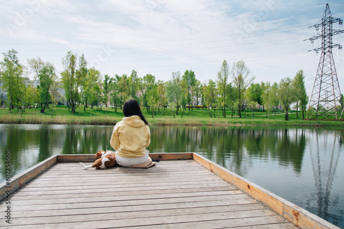 Woman and a dog are sitting on a wooden pier. Woman sitting with her back to the camera, jack russell terrier dog. Beautiful colorful summer natural Idyllic landscape with a lake in the park.