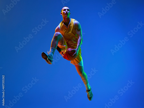 Young muscular man, professional basketball player in motion, training, jumping with ball against blue studio background in neon light. Concept of professional sport, hobby, healthy lifestyle, action