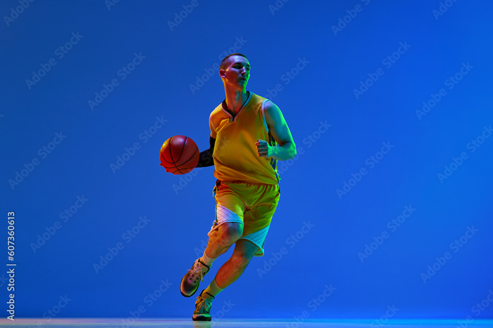 Dynamic image of young man, basketball player in motion during game, training against blue studio background in neon light. Concept of professional sport, hobby, healthy lifestyle, action, game