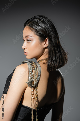 portrait of brunette and asian woman with short hair posing with golden and silver jewelry on shoulder while standing in strapless dress on grey background, wet hairstyle, natural makeup, side view