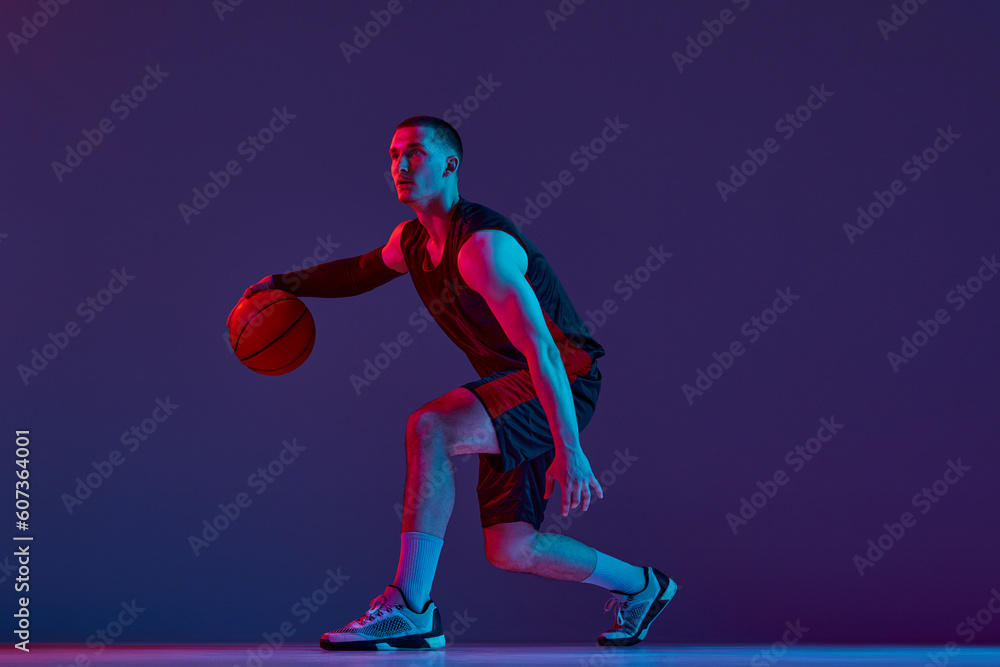 Obraz premium Young muscular man, basketball player in motion, dribbling ball against purple studio background in neon light. Concept of professional sport, hobby, healthy lifestyle, action and motion