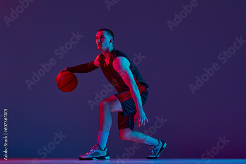 Young muscular man, basketball player in motion, dribbling ball against purple studio background in neon light. Concept of professional sport, hobby, healthy lifestyle, action and motion