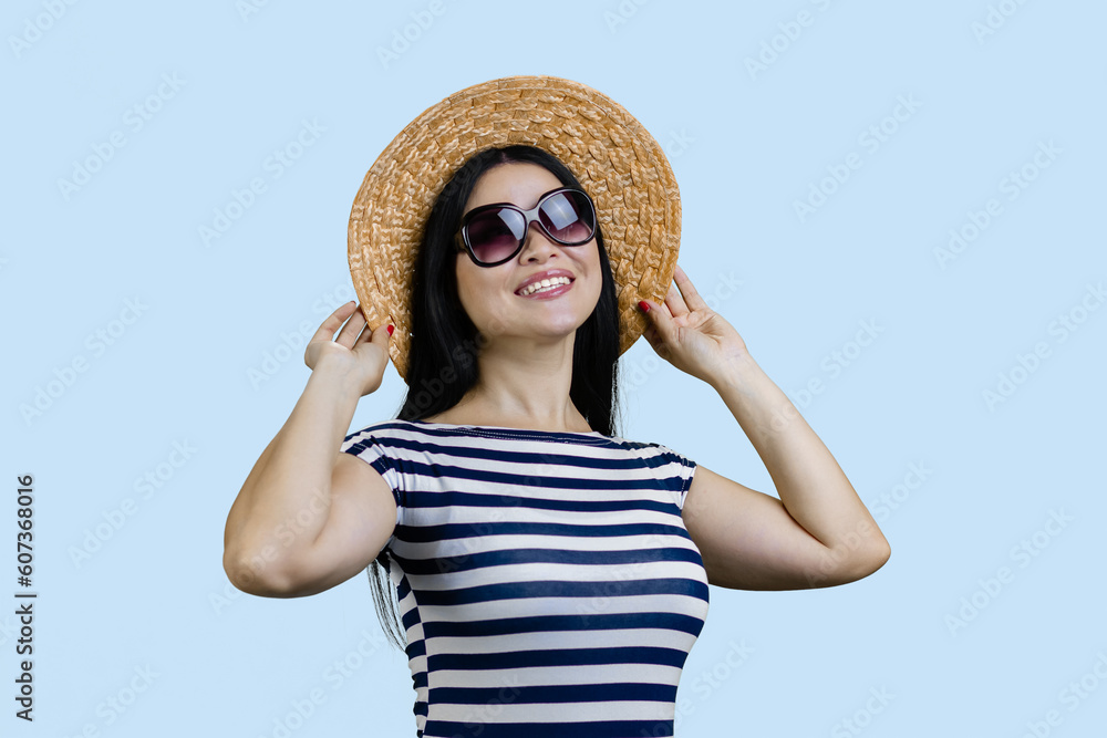 Happy young cheerful brunette woman posing in straw hat and sunglasses. Isolated on pale blue background.