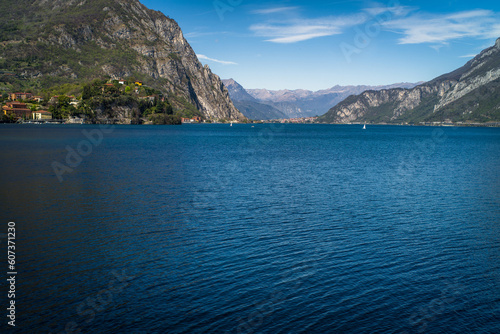 Buildings of the city of Lecco on the shores of Lake Como surrounded by mountains in the background. Sailboats on the waters of the lake and the waterfront.