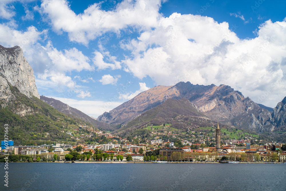 Buildings of the city of Lecco on the shores of Lake Como surrounded by mountains in the background. Sailboats on the waters of the lake and the waterfront.