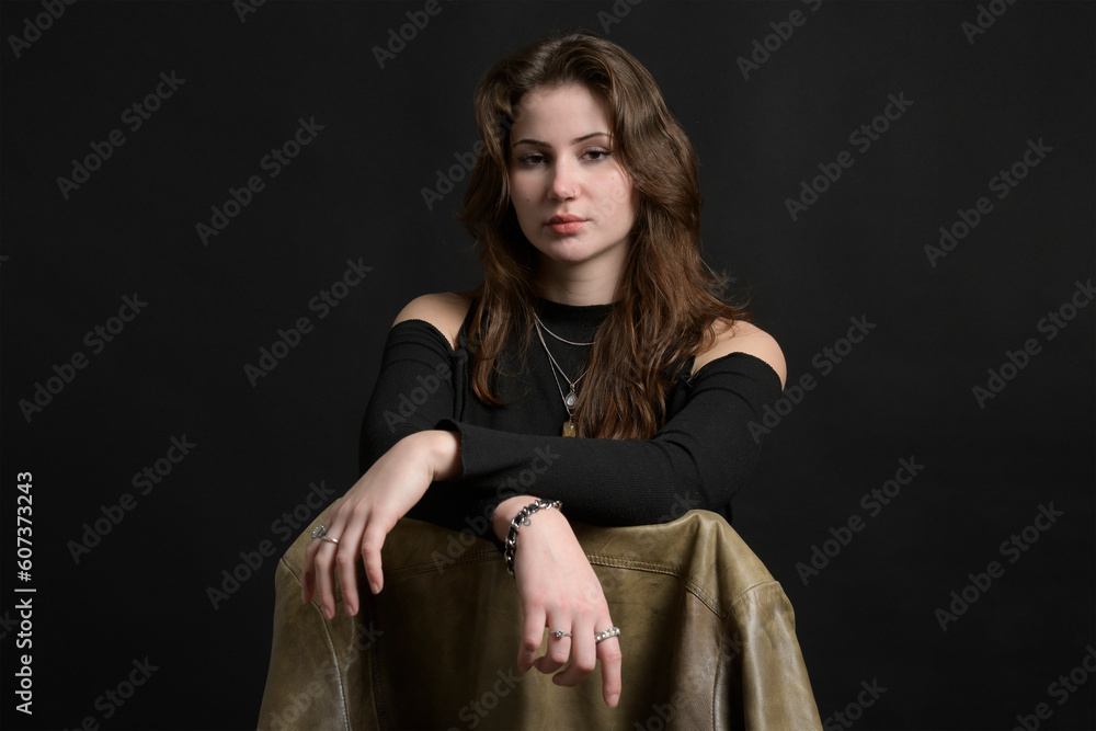Studio portrait of a young lady on a black background sits on a chair with a leather jacket. Caucasian. Brunette. piercing Studio portrait of a young lady on a black background sits on a chair with a leather jacket. Caucasian. Brunette. piercing