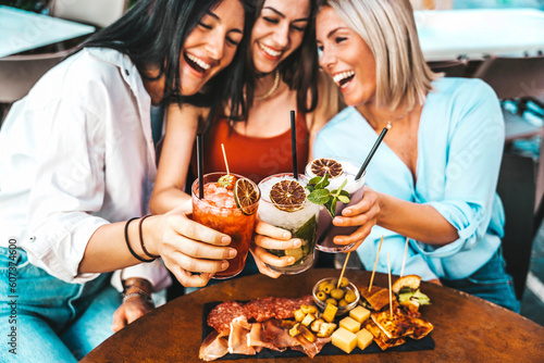Canvas Print Three beautiful young women drinking cocktails sitting at bar restaurant table -