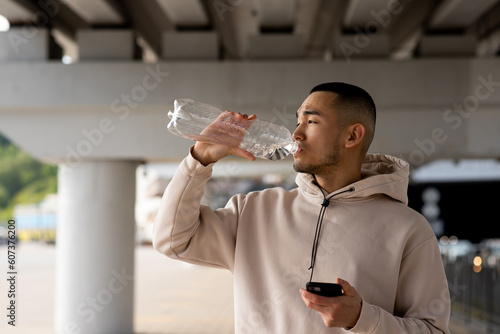 Tired Asian man with smartphone in his hands is drinking water after training outdoors