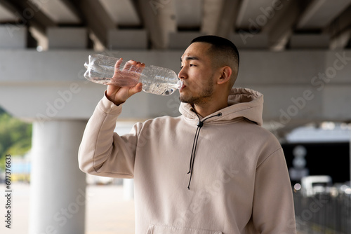 A young guy drinks water from a bottle to quench his thirst after a long run. A young Asian man stands outdoors and drinks water from a bottle.