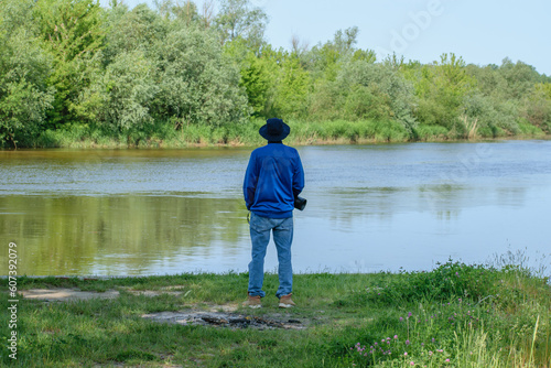 Mężczyzna w kapeluszu ubrany w niebieski strój, stojacy tyłem, patrzący na rzekę 