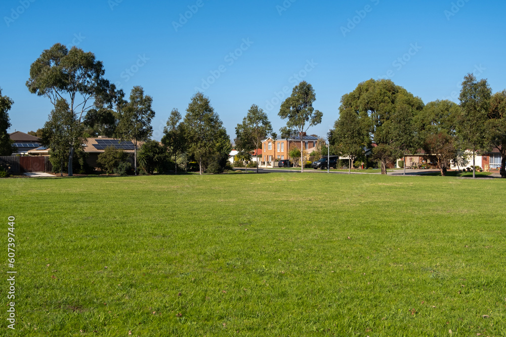 Vacant green grassy sports ground in a public local park in an Australian suburban neighborhood. Background texture of grass lawn with some residential houses in the distance. Vacant green grassy sports ground in a public local park in an Australian suburban neighborhood. Background texture of grass lawn with some residential houses in the distance.