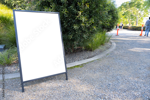Blank white mockup template of a standing board on the side of a path in a park. Empty background texture of advertising easel or information sandwich stand with copy space.