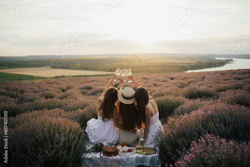 Back view of three young female friends is having picnic with wine, cheese and fresh fruits on lavender field at sunset.	