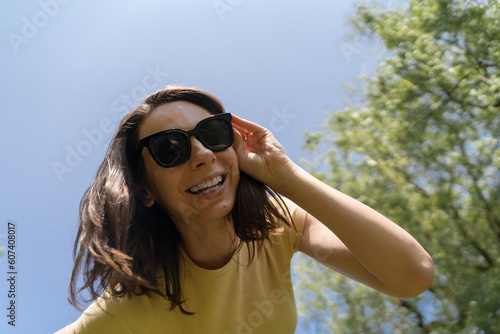 Bottom view of happy millennial woman in yellow t-shirt and sunglasses smiling broadly, looking directly to the camera. © Kseniia