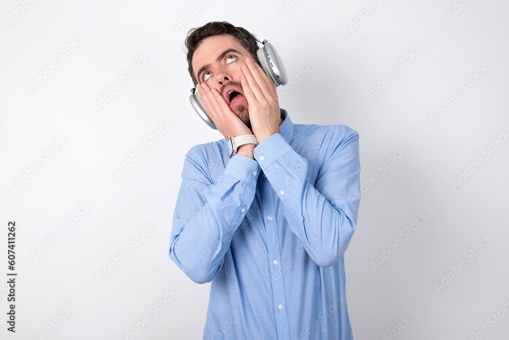 businessman wearing blue t-shirt with headphones over white background keeps hands on cheeks has bored displeased expression. Stressed hopeless model businessman wearing blue t-shirt with headphones over white background keeps hands on cheeks has bored displeased expression. Stressed hopeless model