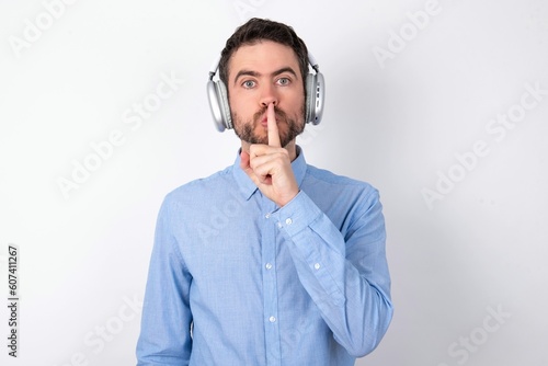 Surprised businessman wearing blue t-shirt with headphones over white background makes silence gesture, keeps finger over lips and looks mysterious at camera photo