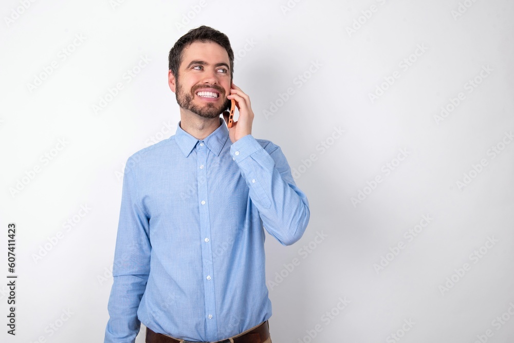 Pleasant looking happy businessman wearing blue t-shirt over white background has nice telephone conversation and looks aside, has nice mood and smiles positively while talks via cell phone Pleasant looking happy businessman wearing blue t-shirt over white background has nice telephone conversation and looks aside, has nice mood and smiles positively while talks via cell phone