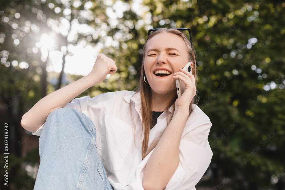 Smiling young girl sitting on ground browsing internet with cellphone wearing casual clothes gesturing like winner. Blonde woman look happy, celebrating win, triumph, success. Girl have a mobile call. Smiling young girl sitting on ground browsing internet with cellphone wearing casual clothes gesturing like winner. Blonde woman look happy, celebrating win, triumph, success. Girl have a mobile call.