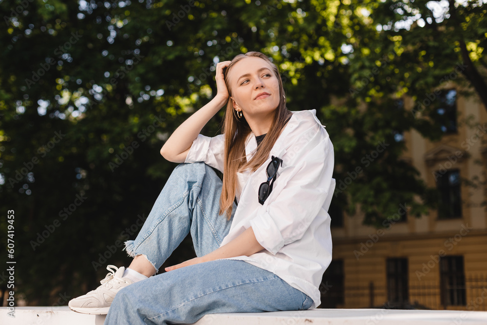 Happy free young woman sitting outdoors look at side on summer park grass. Calm girl enjoy smile and relax in spring city air. Mindset inner light peace concept. Woman wear white shirt and blue jeans. Happy free young woman sitting outdoors look at side on summer park grass. Calm girl enjoy smile and relax in spring city air. Mindset inner light peace concept. Woman wear white shirt and blue jeans.