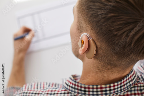 Close up shot of man with hearing aid drawing on paper. View from behind of hearing impaired young man working at desk in office or home
