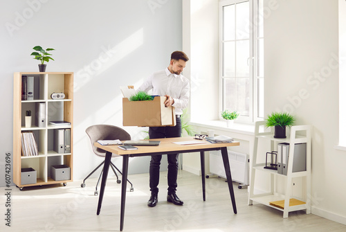 Fired male employee emptying his desk, making room for new worker. Young man standing by his former working desk in office and putting his belongings such as folders and green plants in cardboard box © Studio Romantic