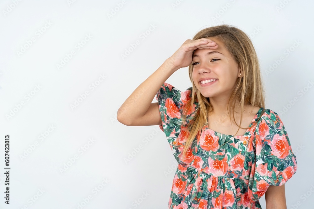 beautiful teen girl wearing flowered dress over white studio background very happy and smiling looking far away with hand over head. Searching concept.