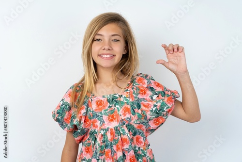 beautiful teen girl wearing flowered dress over white studio background smiling and confident gesturing with hand doing small size sign with fingers looking and the camera. Measure concept