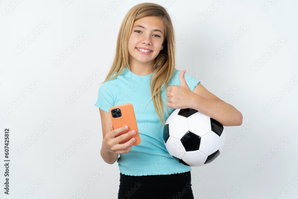 Portrait of beautiful caucasian teen girl wearing sportswear holding a football ball using and texting with smartphone  happy with big smile doing ok sign, thumb up with fingers, excellent sign