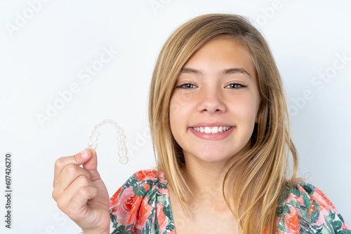 beautiful caucasian teen girl wearing flowered blouse over white wall holding an invisible braces aligner, recommending this new treatment. Dental healthcare concept.
