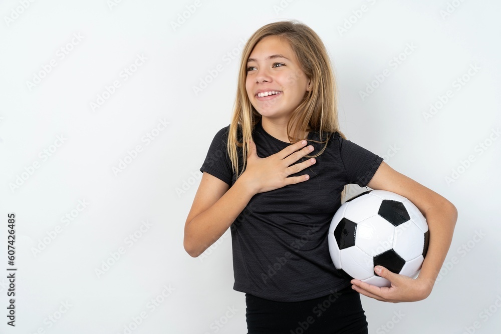 Joyful beautiful caucasian teen girl wearing sportswear holding a football ball over white wall expresses positive emotions recalls something funny keeps hand on chest and giggles happily. Joyful beautiful caucasian teen girl wearing sportswear holding a football ball over white wall expresses positive emotions recalls something funny keeps hand on chest and giggles happily.