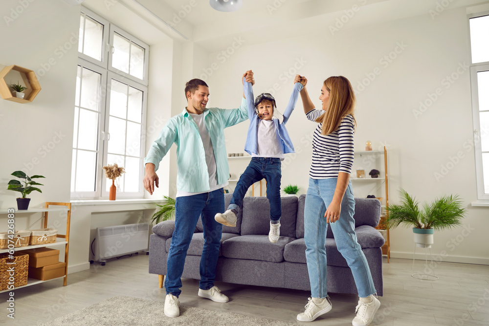 Happy smiling family having fun with their child son in the living room at home. Couple lifting in the air their laughing cheerful boy kid in pilot's glasses and playing plane with him. Happy smiling family having fun with their child son in the living room at home. Couple lifting in the air their laughing cheerful boy kid in pilot's glasses and playing plane with him.