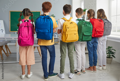 Back to school concept. Group of school children with backpacks. Backside view from behind six students with colorful pink, blue, yellow, green, red and grey bags standing together in the classroom
