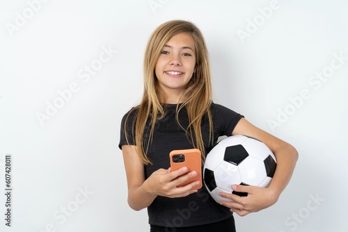 beautiful caucasian teen girl wearing sportswear  over white wall holds mobile phone in hands and rejoices positive news  uses modern cellular