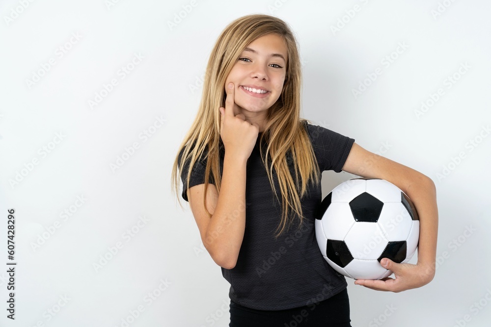 Happy beautiful caucasian teen girl wearing sportswear over white wall with toothy smile, keeps index fingers near mouth, fingers pointing and forcing cheerful smile Happy beautiful caucasian teen girl wearing sportswear over white wall with toothy smile, keeps index fingers near mouth, fingers pointing and forcing cheerful smile