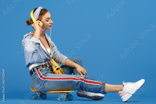 Full length photo of carefree young woman with yellow headphones sitting on skateboard over blue background.