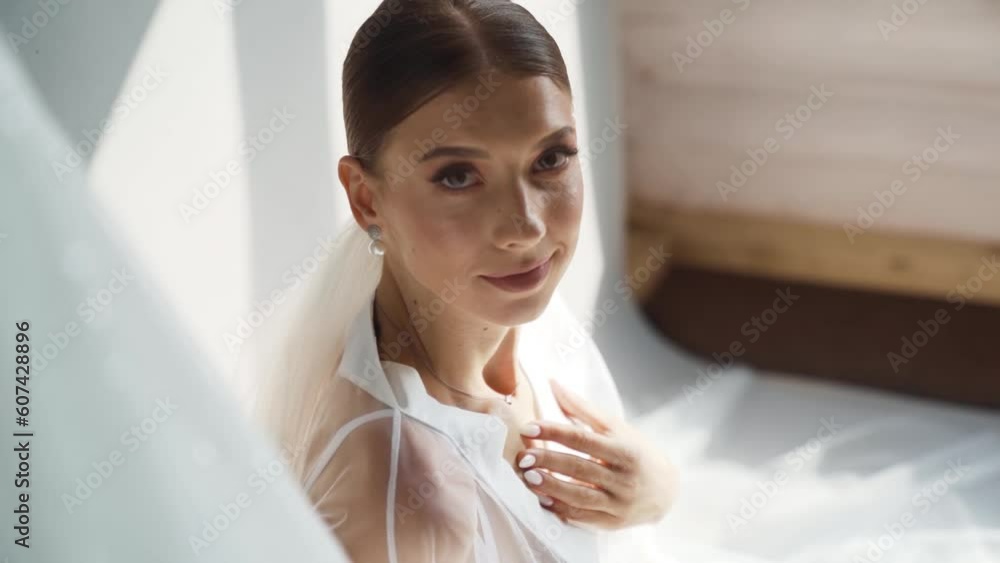 Elegant bride posing near hanging wedding dress the morning of the wedding day Elegant bride posing near hanging wedding dress the morning of the wedding day