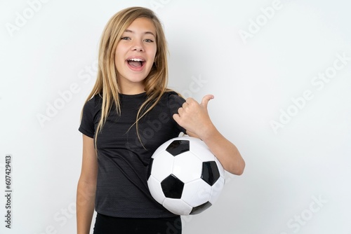 Impressed beautiful caucasian teen girl wearing sportswear and football ball over white wall point back empty space © Jihan