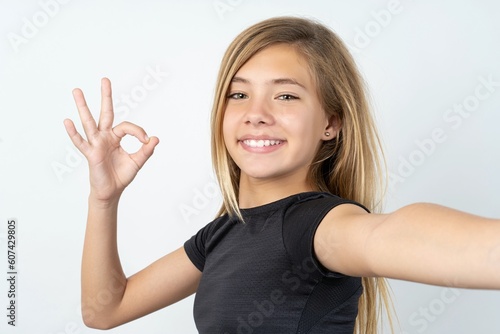 Portrait of pretty cheerful beautiful caucasian teen girl wearing sportswear over white wall make selfie show okey symbol