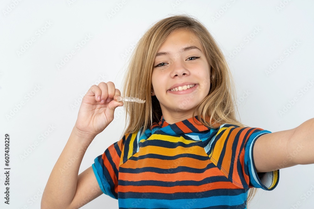 beautiful caucasian teen girl wearing striped T-shirt over white wall make selfie holding an invisible braces aligner, recommending this new treatment. Dental healthcare concept. beautiful caucasian teen girl wearing striped T-shirt over white wall make selfie holding an invisible braces aligner, recommending this new treatment. Dental healthcare concept.