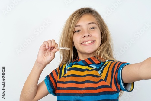 beautiful caucasian teen girl wearing striped T-shirt over white wall make selfie holding an invisible braces aligner, recommending this new treatment. Dental healthcare concept.
