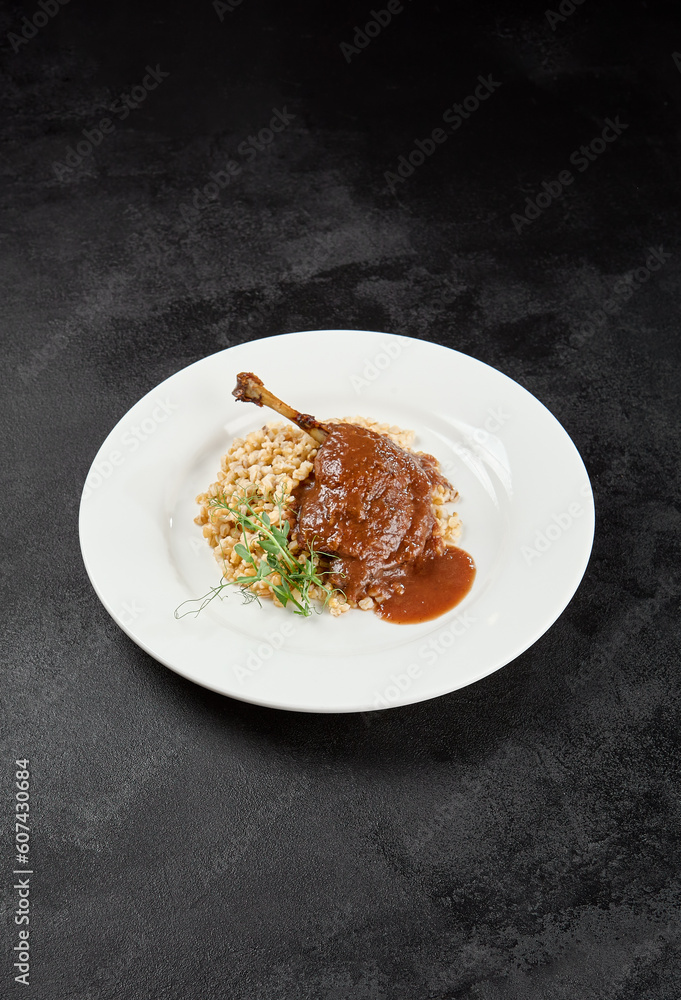 Classic dish french cuisine - roasted duck leg in porto sauce with garnish on black background. Cooked duck leg with pearl barley on white plate on dark concrete table. Duck confit in minimal style.