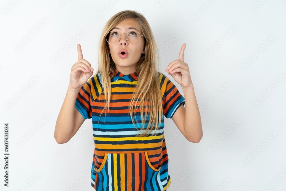 beautiful caucasian teen girl wearing striped T-shirt over white wall being amazed and surprised looking and pointing up with fingers showing something strange. beautiful caucasian teen girl wearing striped T-shirt over white wall being amazed and surprised looking and pointing up with fingers showing something strange.