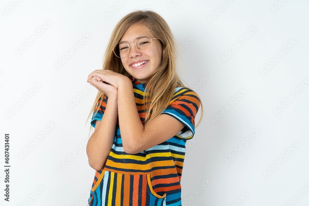 Dreamy beautiful caucasian teen girl wearing striped T-shirt over white wall with pleasant expression, closes eyes, keeps hands crossed near face, thinks about something pleasant